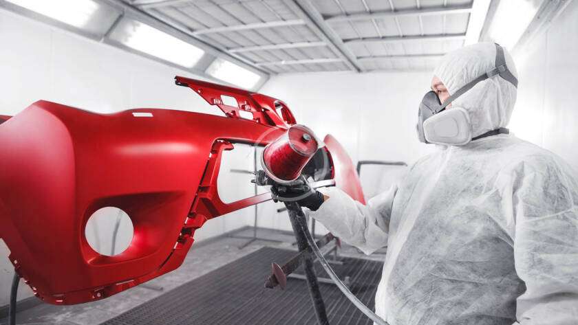 Person spray painting a red car bumper in a workshop.