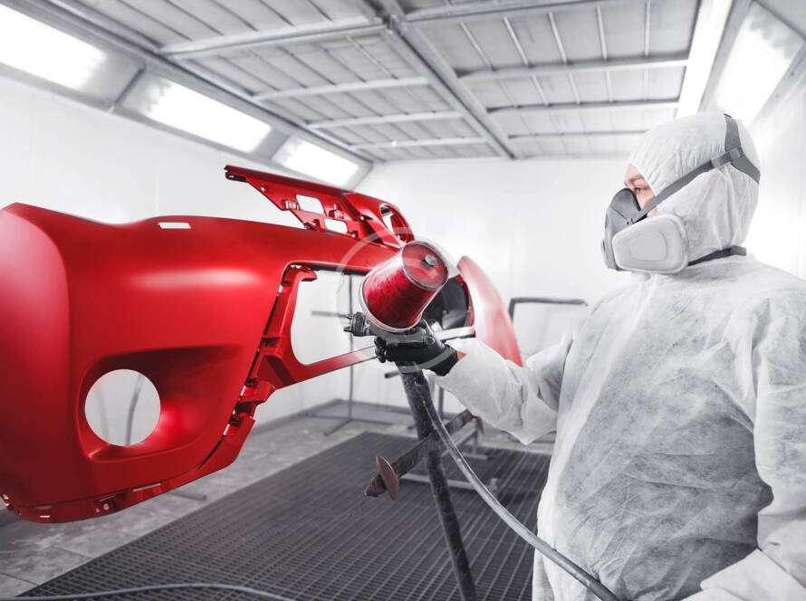 Person spray painting a red car bumper in a workshop.