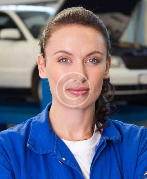 A confident woman mechanic in blue overalls standing in a garage.