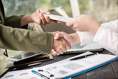 Two people shaking hands and exchanging documents in a business setting.